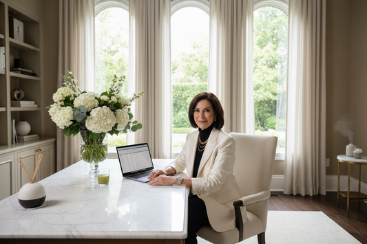 Polished woman in her 50s at a marble desk in a luxurious home office with laptop, fresh flowers, wearing a tailored blazer and elegant jewelry, soft natural light, confident focused expression