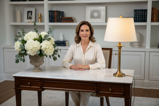 Elegant woman in her 50s sitting composed at a luxurious home desk, soft warm lighting, silk blouse, tasteful jewelry, fresh flowers, marble surface, radiant and confident